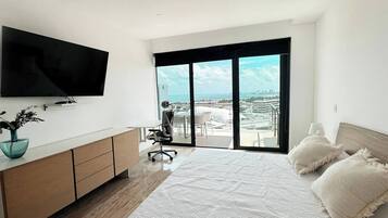 Une chambre moderne avec un grand lit, un téléviseur fixé au mur, un bureau avec une chaise et un balcon offrant une vue sur la ville et l’océan.