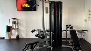 Un gimnasio con una estructura para sentadillas, banca de pesas y una televisión mostrando 'MWAKI'.