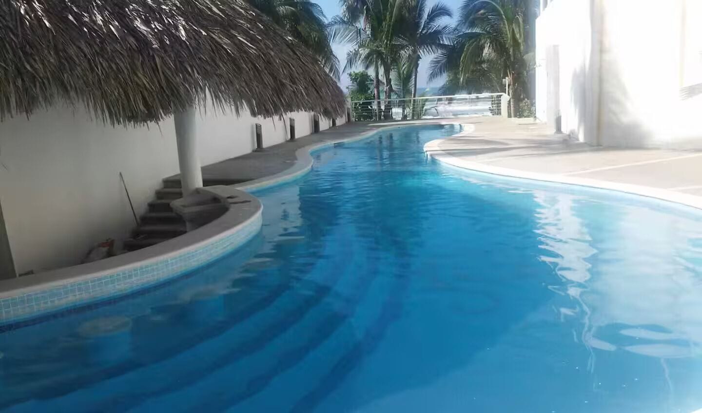 A swimming pool with a curved design, surrounded by a white wall and a thatched roof structure.