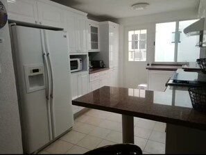 A kitchen with white cabinets, a refrigerator, a microwave, and a window with blinds. - Villa Brisas del Mar - Acapulco Diamante, Club de Playa (Acapulco de Juarez)