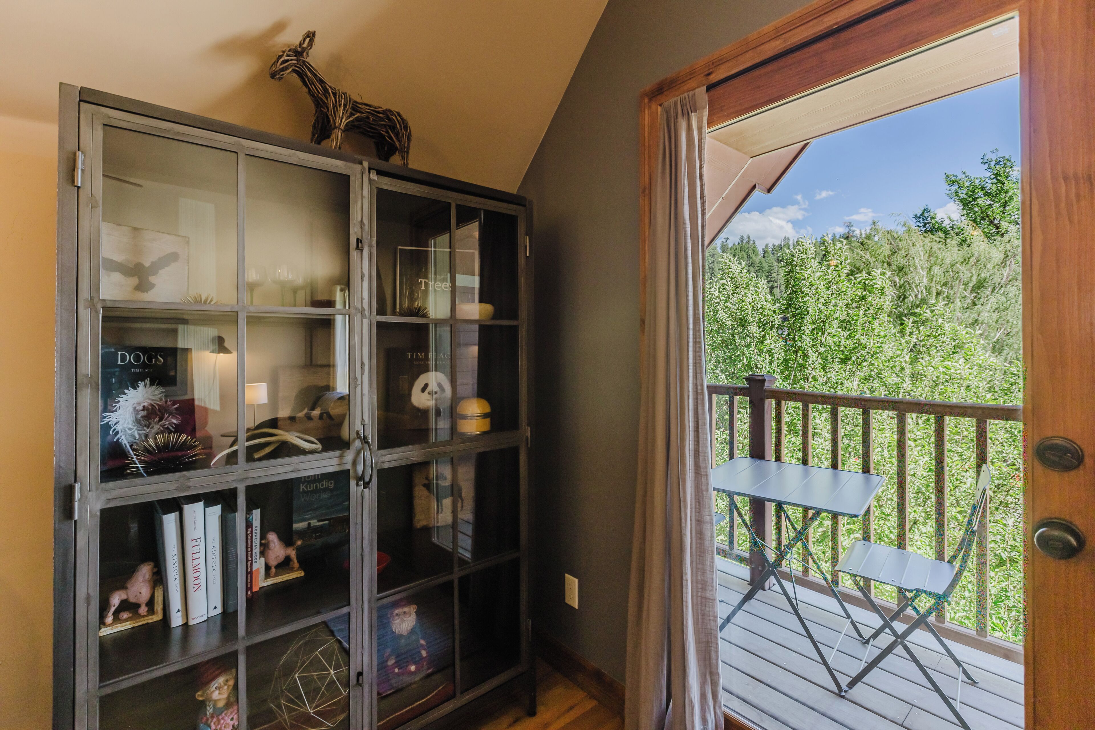 A room with a glass-fronted bookcase, a small table, and a view of a wooden deck with a railing.