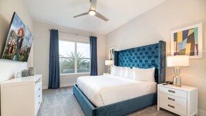 Un dormitorio con cama, mesitas de noche, ventilador de techo, ventana con cortinas y obras de arte en las paredes.