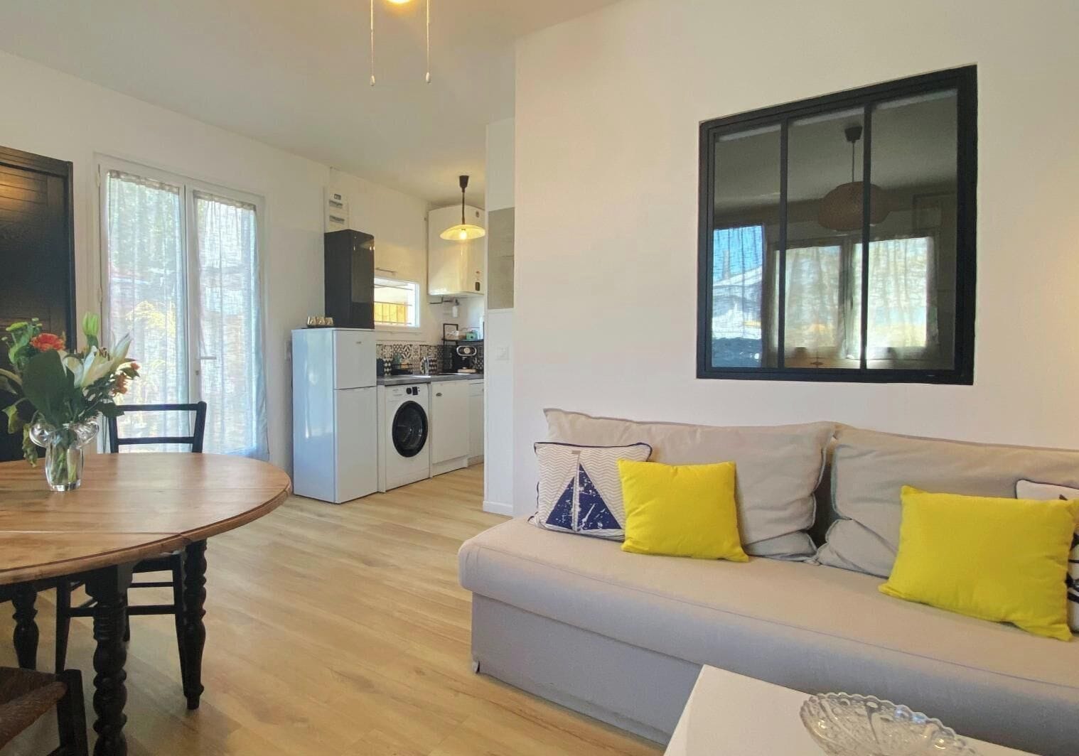 A modern apartment with a dining area, a sofa with yellow cushions, a kitchen with a washing machine, and a large window.
