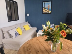 Ein Wohnzimmer mit einer Couch, einem runden Holztisch mit Blumen in einer Vase, einer Wandleuchte und einem gerahmten Bild.