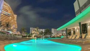 Un espace piscine avec des chaises longues, un grand bâtiment aux fenêtres illuminées et un ciel nocturne dégagé.