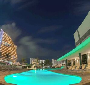 Zona de piscina con sillones de descanso, un edificio grande con ventanas iluminadas y cielo nocturno despejado.