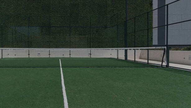 Un court de tennis doté d’une surface verte et de lignes blanches délimitant le terrain, entouré d’une clôture et bordé d’un bâtiment dont le mur est vert.
