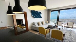 Une chambre d’hôtel moderne dotée d’une grande fenêtre, d’un canapé, de chaises jaunes, d’une table à manger et de luminaires suspendus.