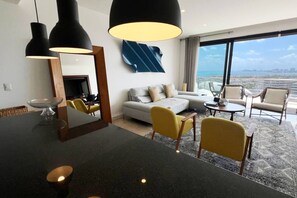 Una habitación de hotel moderna con un ventanal grande, un sofá, sillas amarillas, una mesa de comedor e iluminación colgante.