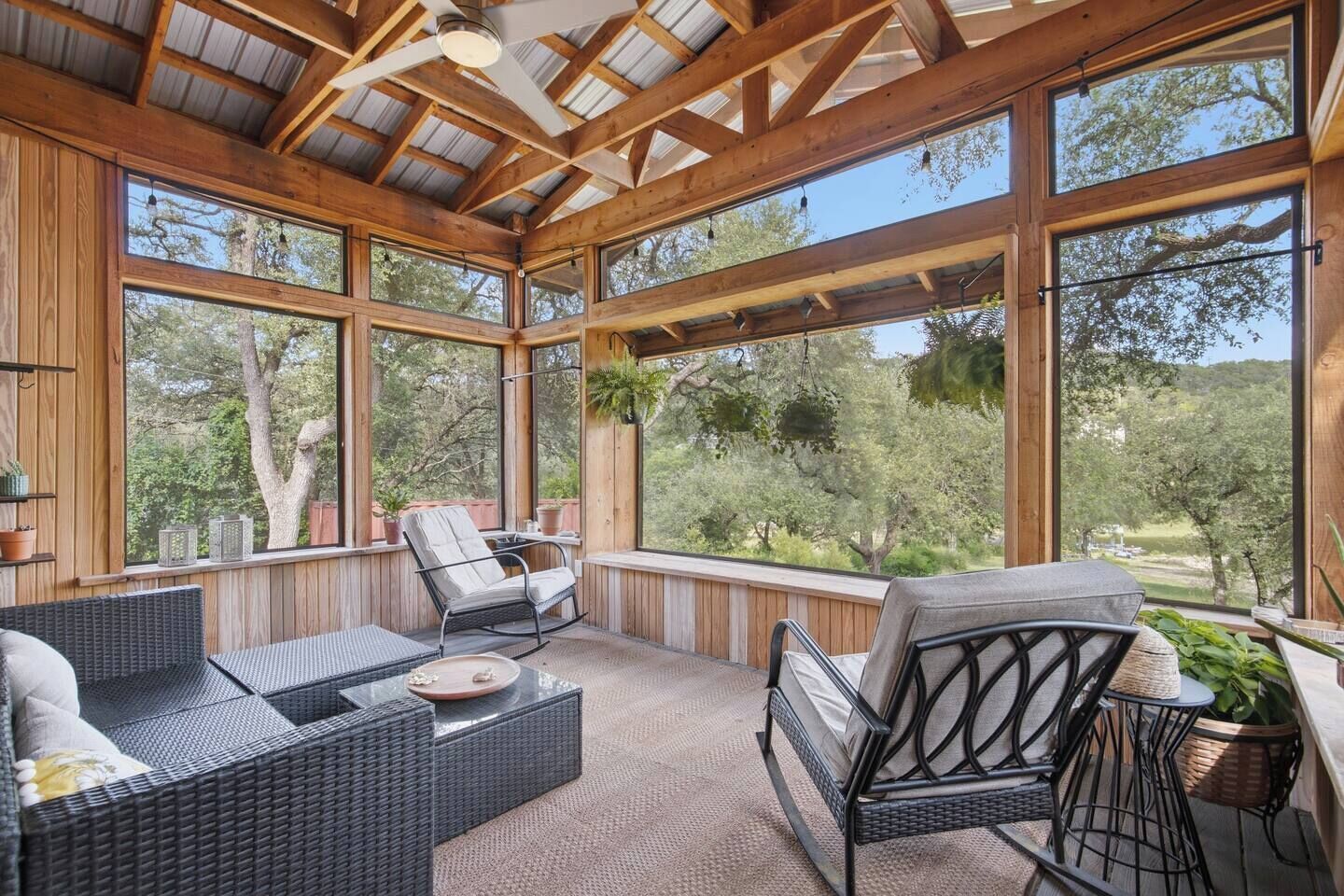 A covered outdoor seating area with wicker furniture, large windows, and a view of trees.