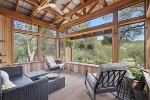 Un espacio de estar al aire libre cubierto con mobiliario de mimbre, ventanas amplias y vistas a árboles.