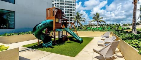 A playground with a slide and climbing structure, surrounded by outdoor seating and palm trees.