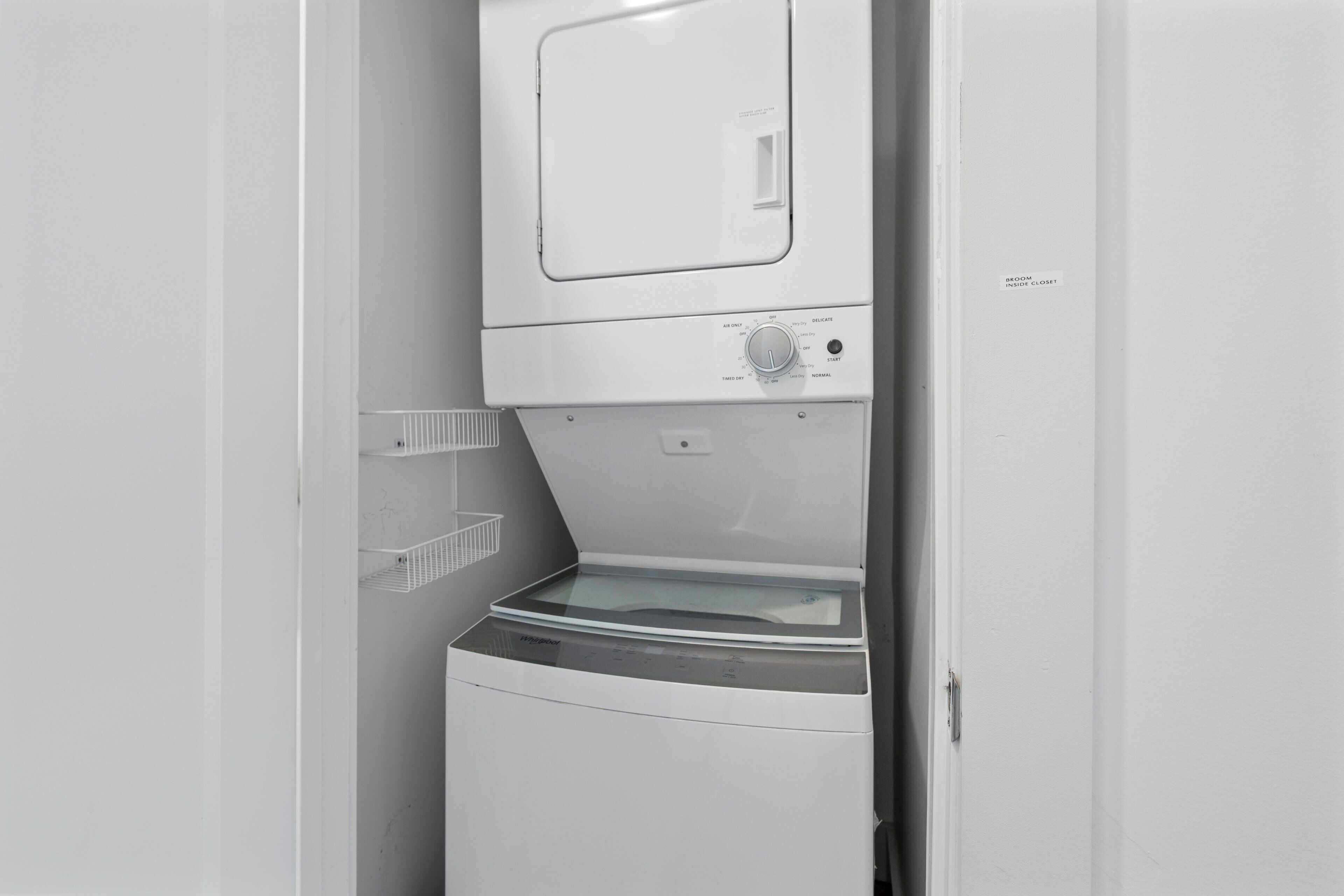 A compact, white laundry room with a front-loading washer and dryer, a small shelf, and a door with a label.