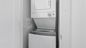 Uma lavanderia compacta e branca, com máquina de lavar e secar de abertura frontal, uma prateleira pequena e uma porta com etiqueta.