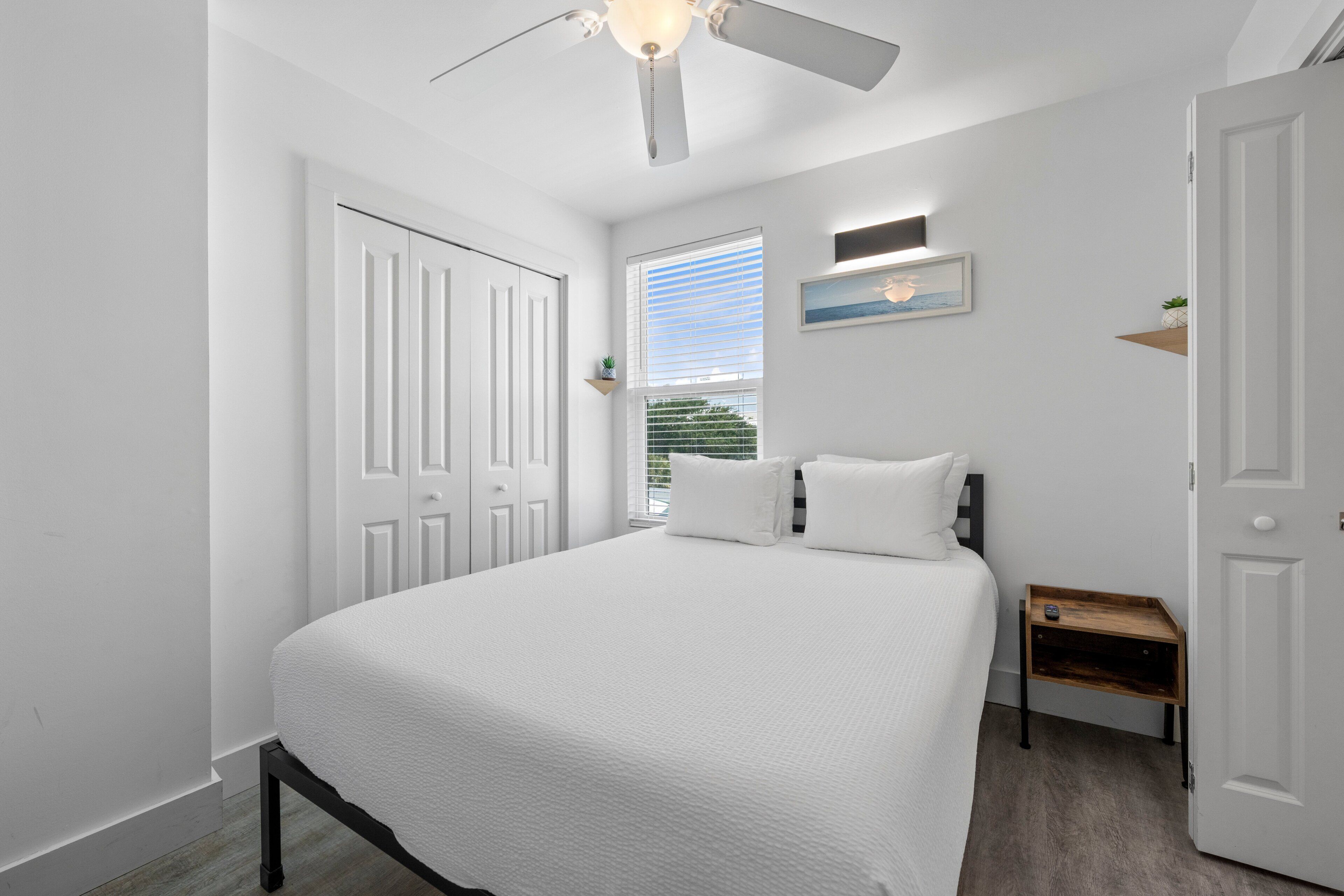 Ein Schlafzimmer mit Bett, Deckenventilator, Fenster mit Jalousien, Nachttisch und Tür.