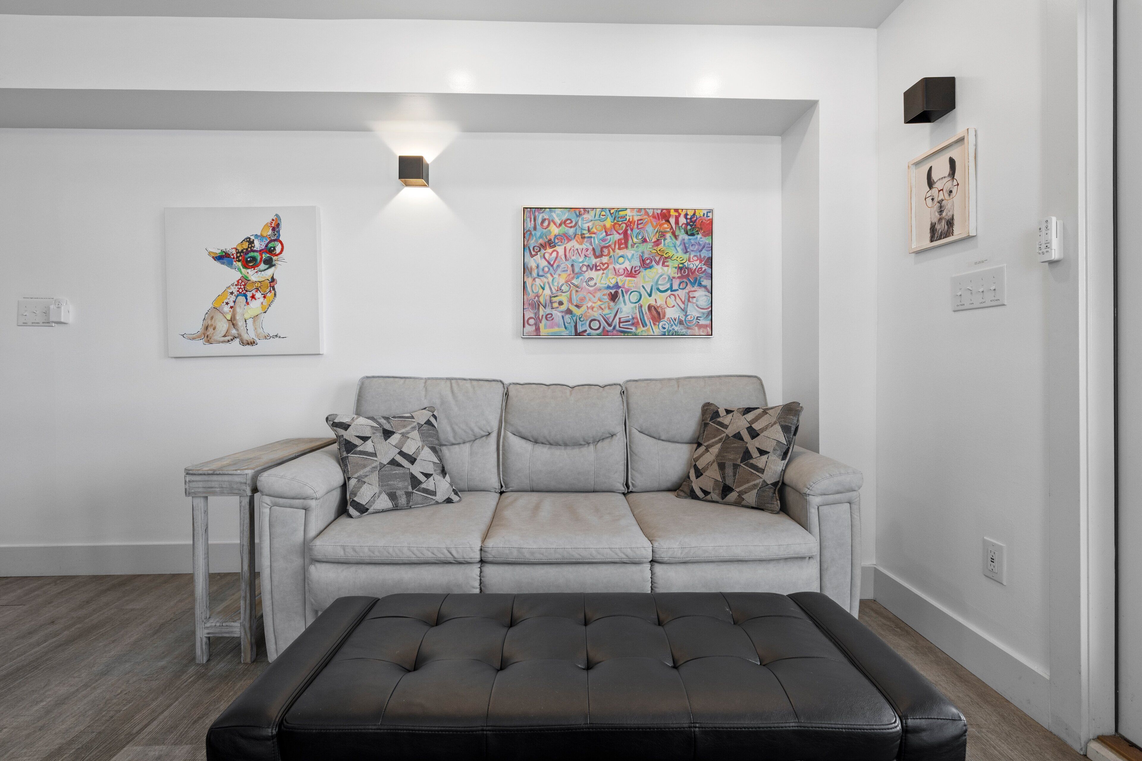 Een moderne woonkamer met een grijze bank, een zwart voetenbankje en twee kunstwerken aan de muur.