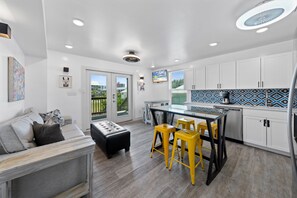 A modern kitchen with white cabinets, a dining area with yellow stools, and a large window overlooking a green outdoor area. - Pipedreams West (Gulf Shores)