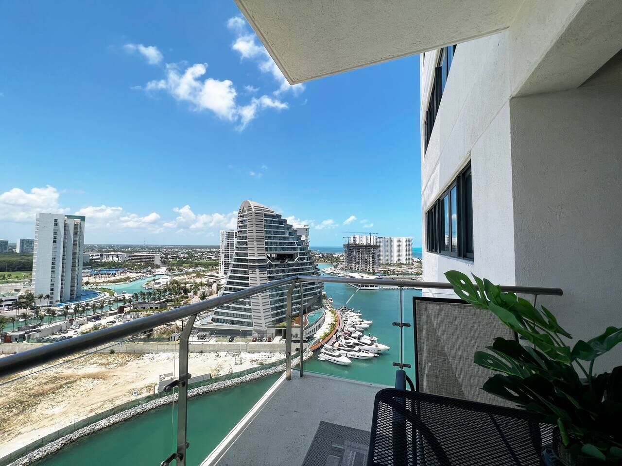 Un balcón con vista a un puerto deportivo, un edificio moderno y otras construcciones a lo lejos.