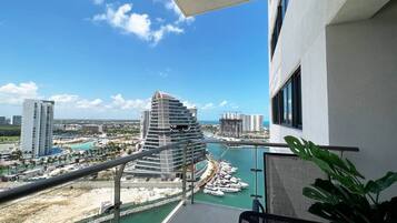 Un balcón con vista a un puerto deportivo, un edificio moderno y otras construcciones a lo lejos.