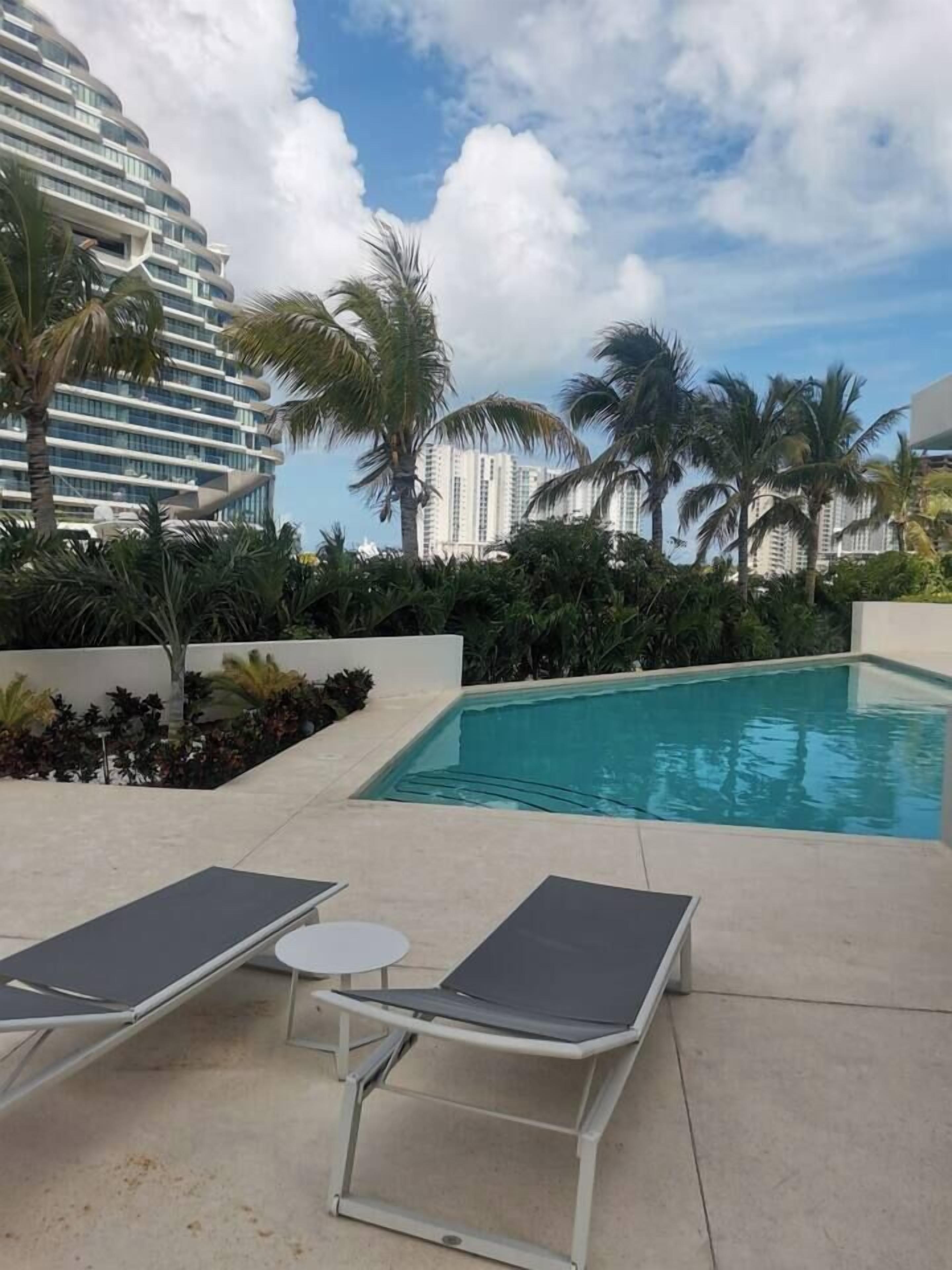 Une piscine entourée de palmiers, avec des chaises longues et une petite table, et un bâtiment visible au loin.