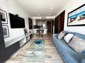 Una sala de estar moderna con un sofá azul, una mesa de centro de vidrio y una cocina al fondo.