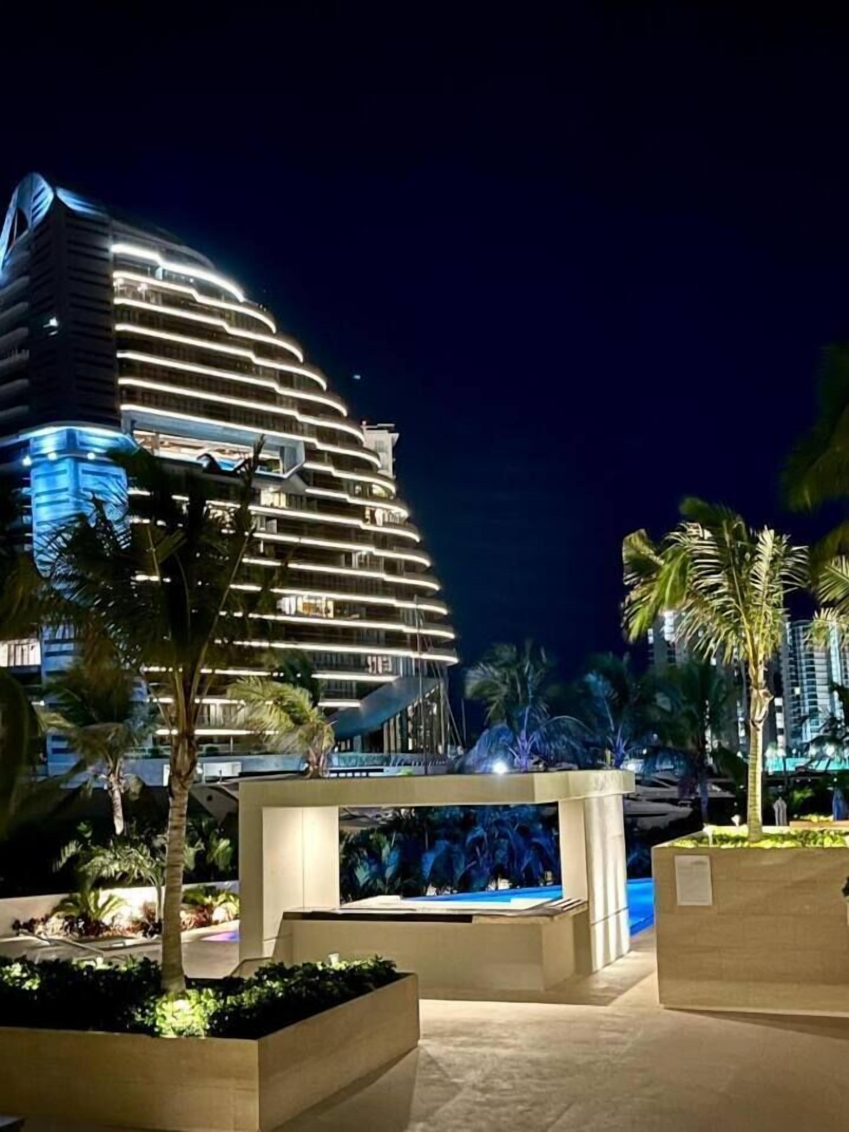 Ein modernes Hotel mit geschwungener Fassade, einem Poolbereich mit Wasserspiel und Palmen.