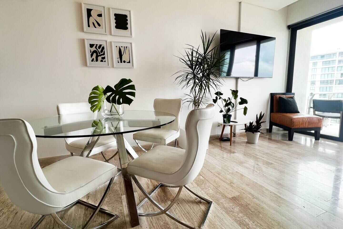 Un salon moderne avec une table en verre entourée de chaises, un téléviseur à écran plat fixé au mur et plusieurs plantes en pot.