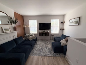 Ein modernes Wohnzimmer mit dunklen Sofas, einem Flachbildfernseher und einem großen Fenster mit Jalousien.