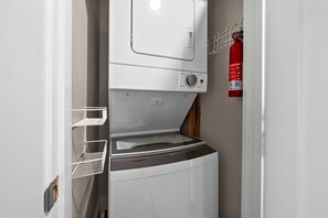 A compact washer and dryer unit with a control panel, a metal shelf, and a fire extinguisher mounted on the wall. - Pipedreams East (Gulf Shores)