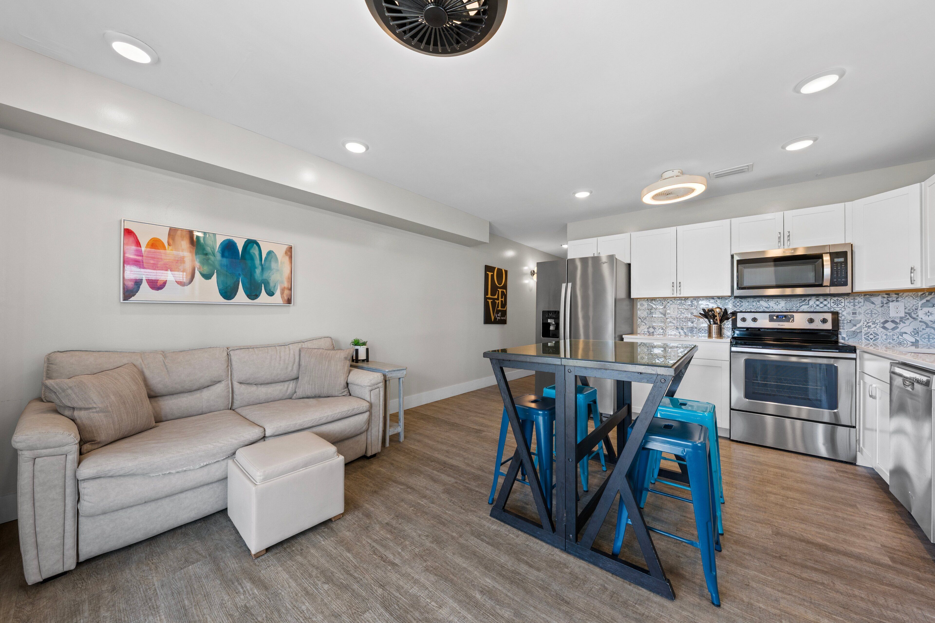 A modern kitchen with stainless steel appliances, a dining area with blue stools, and a beige sofa.
