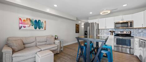 A modern kitchen with stainless steel appliances, a dining area with blue stools, and a beige sofa.