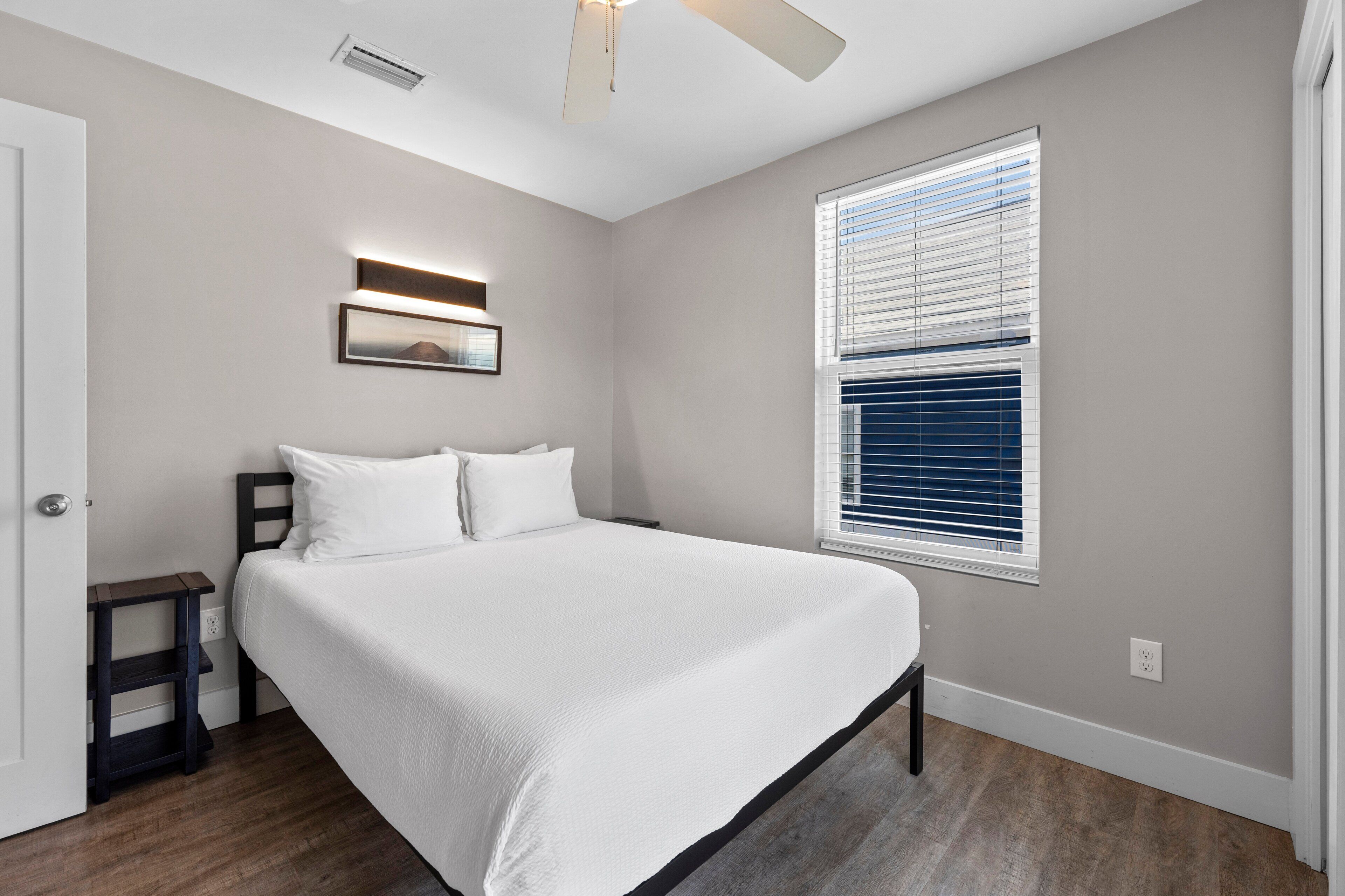 Une chambre à coucher comprenant un lit, une table de chevet, un ventilateur de plafond, une fenêtre avec des stores et un luminaire fixé au mur.