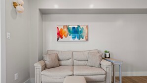 En soffa i en neutral färgskala med en tillhörande pall, ett litet sidobord med en växt och en färgstark, abstrakt tavla på väggen.