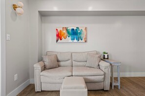 A neutral-toned sofa with a matching ottoman, a small side table with a plant, and a colorful abstract painting on the wall. - Pipedreams East (Gulf Shores)