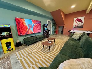 A living room with a large flat-screen TV, a gaming console, a sofa, a coffee table, and a patterned rug. - Hot Tub · Arcade · Sleeps 11 · Walk Downtown/Beach (Ocean Shores)
