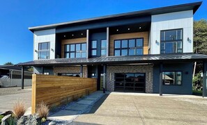 A modern two-story building with a garage, large windows, and a covered entrance area. - 2 Houses · Arcade+Speakeasy · Sleep 20 · 100" TV (Ocean Shores)