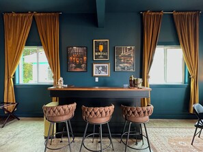 A bar area with a wooden counter and three bar stools, framed pictures on the wall, and curtains on the windows. - 2 Houses · Arcade · Speakeasy · Sauna · Sleeps 20 (Ocean Shores)