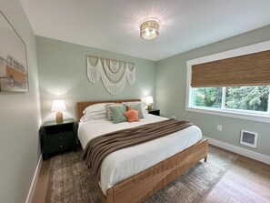 A bedroom with a bed, two bedside tables, a wall-mounted macrame piece, a window with a blind, and a ceiling light fixture. - 2 Houses · Arcade+Speakeasy · Sleep 20 · 100" TV (Ocean Shores)