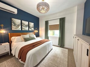 A bedroom with a bed, bedside tables, a wall-mounted air conditioner, a chandelier, and a window with curtains. - Hot Tub+Speakeasy · Sleeps 9 · Walk Downtown/Beach (Ocean Shores)