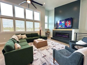 Een woonkamer met een hoekbank, een open haard, een flatscreen televisie en grote ramen.