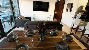 Een eetruimte met een houten tafel voor zes personen, een flatscreen televisie erboven en uitzicht op buiten door grote ramen.
