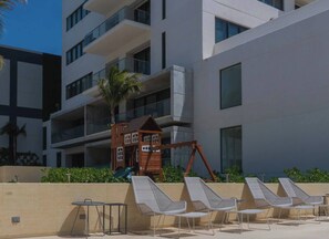 Un edificio moderno con un parque infantil, sillas de descanso y una zona de piscina.