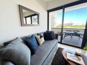 Una sala de estar moderna con un sofá gris, una mesa de centro redonda y un ventanal con vistas a un balcón y edificios.