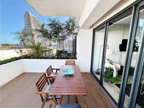 A wooden dining table with chairs, a glass balustrade, and a view of a modern building and palm trees. - Desayuna en tu terraza · Disfruta las vistas · TOP (Cancún)