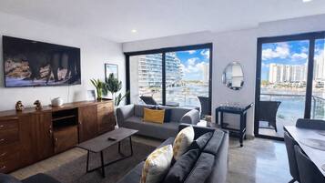 Un salon moderne avec un canapé, une table basse et un téléviseur. Il y a un balcon avec vue sur la ville.