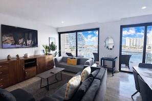 Una sala de estar moderna con sofá, mesa de centro y televisor. Hay un balcón con vistas a la ciudad.