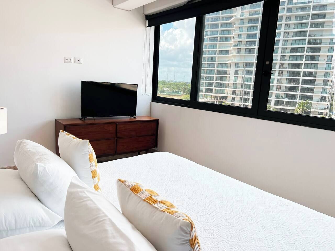 Une chambre d’hôtel avec un lit, un téléviseur posé sur une commode en bois et une fenêtre donnant sur un immeuble.