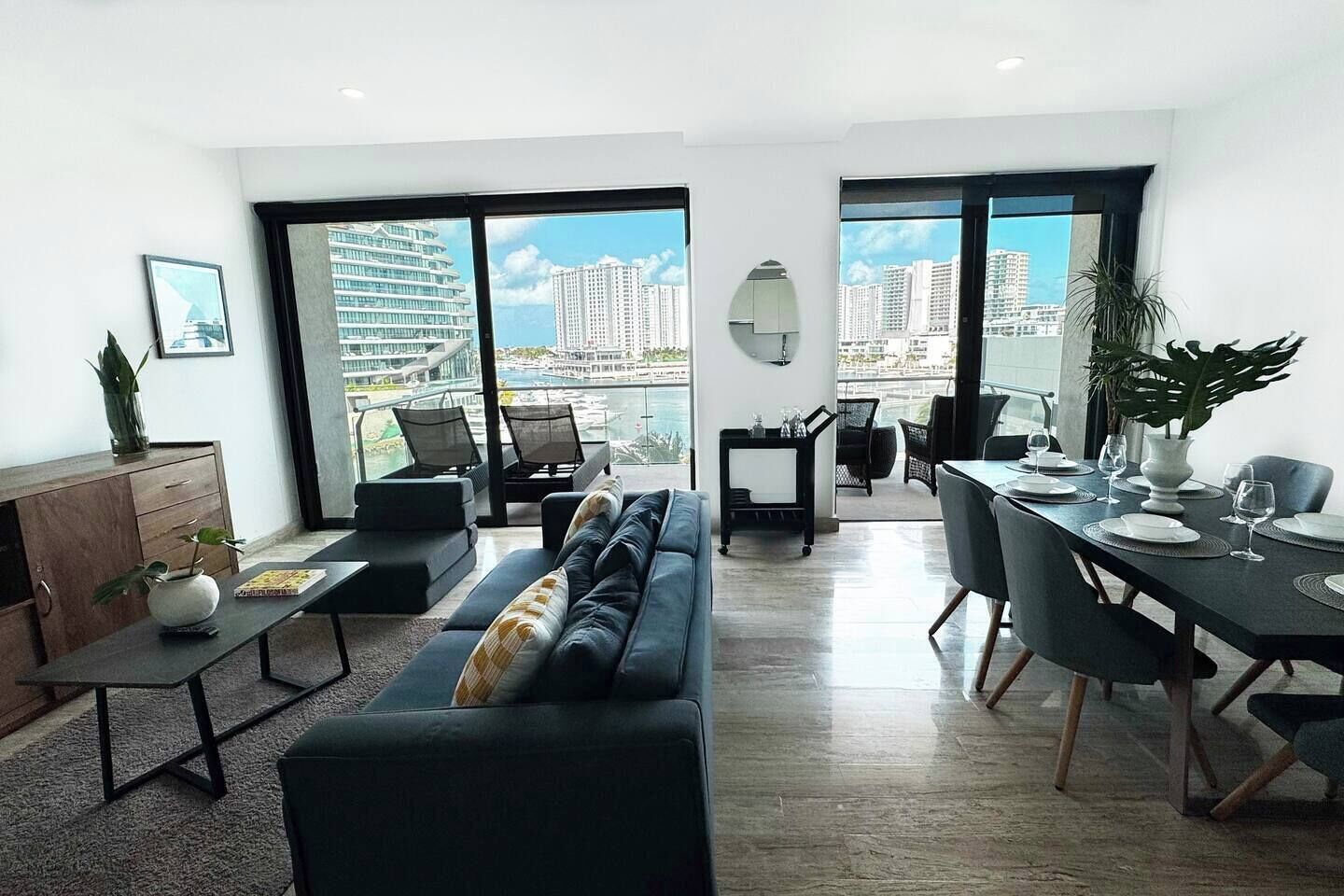 Un salon moderne comprenant un canapé, une table à manger et des chaises, de grandes fenêtres offrant une vue sur la ville, et une petite plante posée sur la table d’appoint.