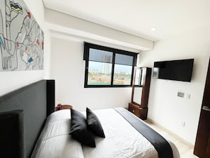 Un dormitorio con cama, ventana, televisor de pantalla plana y una obra de arte en la pared.