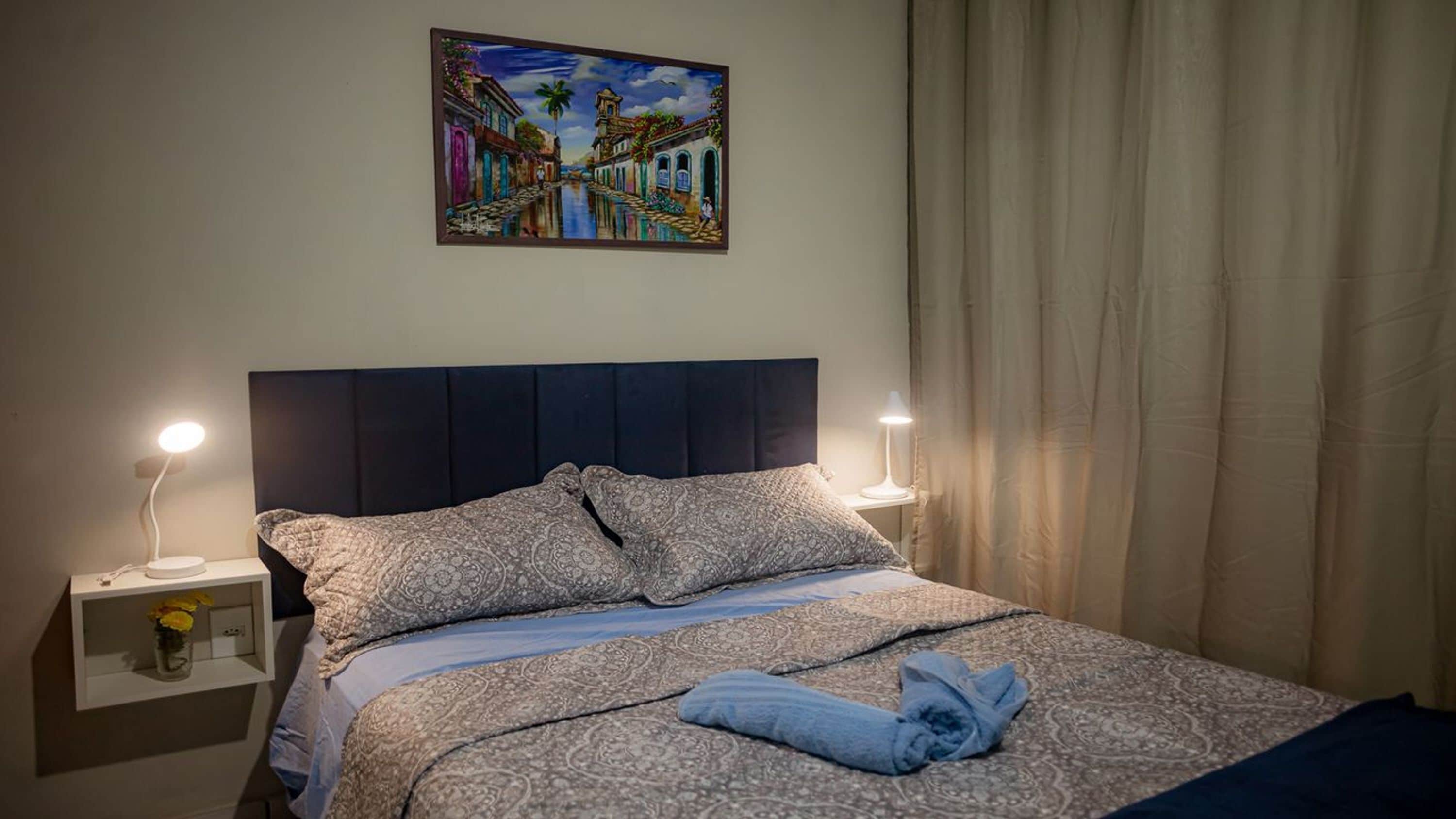 Una cama bien hecha con ropa de cama estampada, dos lámparas de noche, una mesita de noche con un jarrón de flores y un cuadro de una escena callejera sobre la cama.